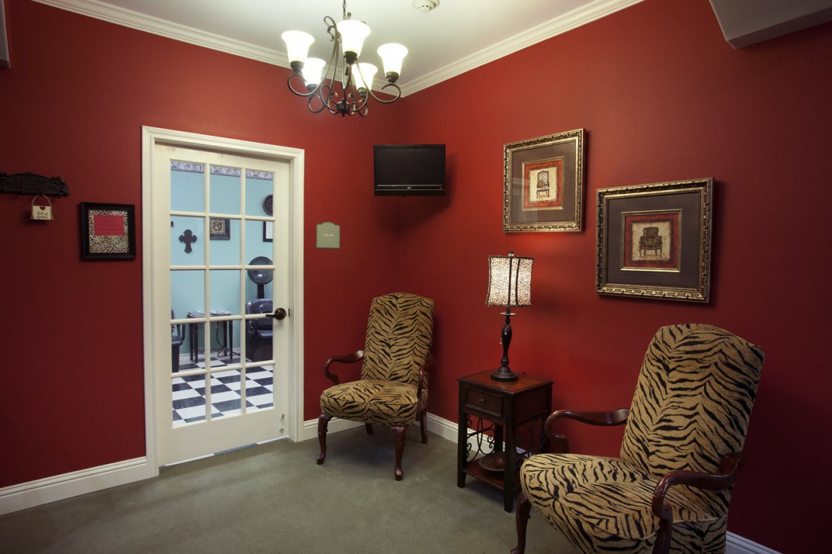A cozy waiting room with red walls, featuring two patterned chairs, a small table with a lamp, and wall art.