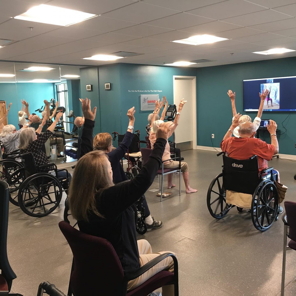 A group of seniors participating in an exercise class, with some in wheelchairs, engaging in an indoor physical activity session.