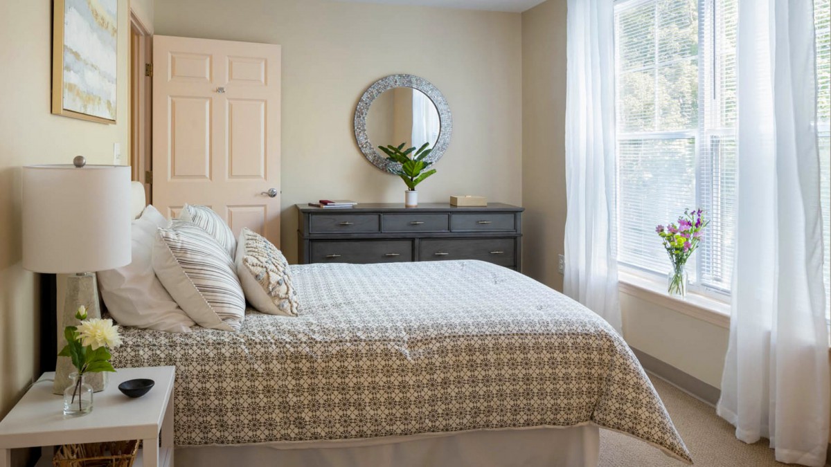 A cozy bedroom with a neatly made bed, dresser, and round mirror by the window.