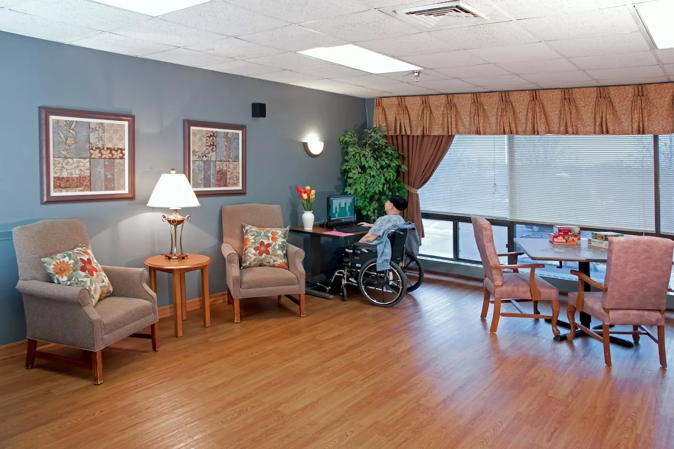 A cozy lounge area in a nursing home with comfortable chairs, a wheelchair user by a desk, and large windows letting in natural light.