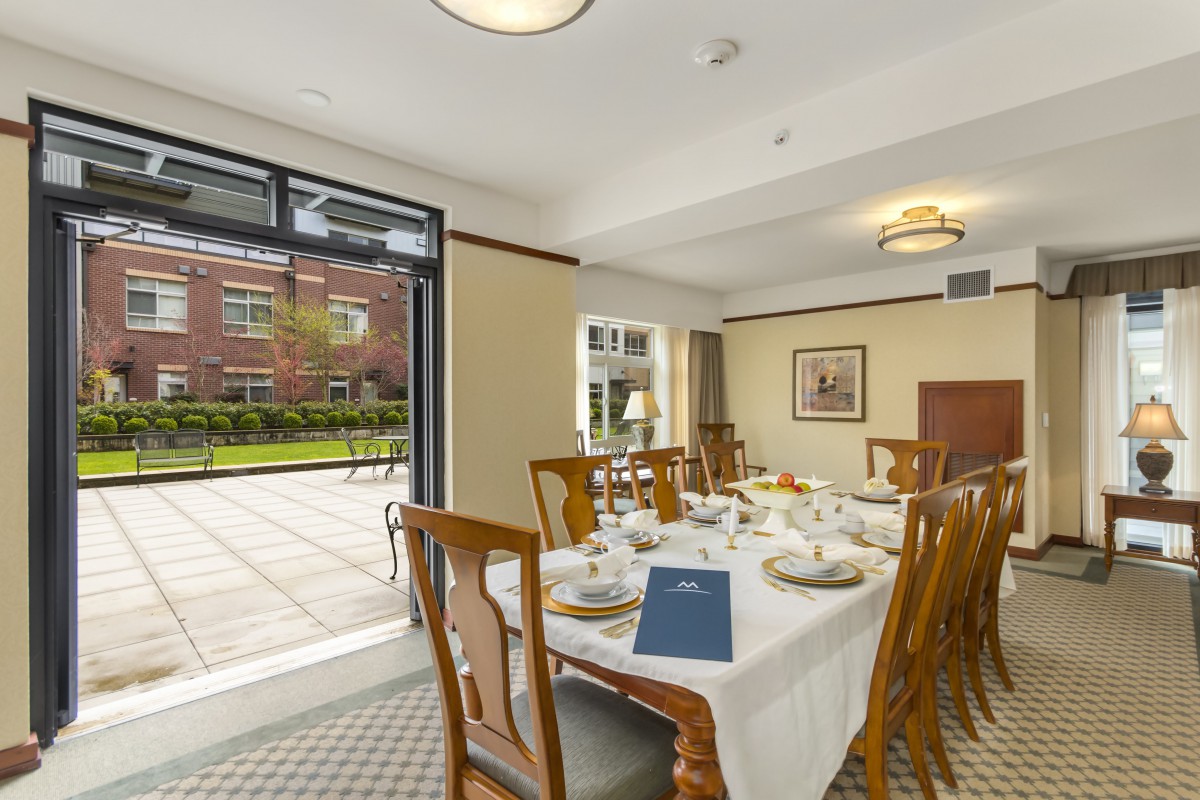 A well-lit dining room with a table set for a meal, featuring wooden chairs and windows overlooking a garden.