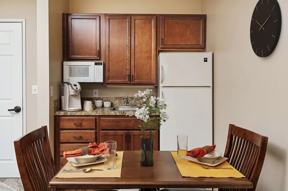 A cozy kitchen with wooden cabinets and a dining table set for two, featuring a white refrigerator and microwave.