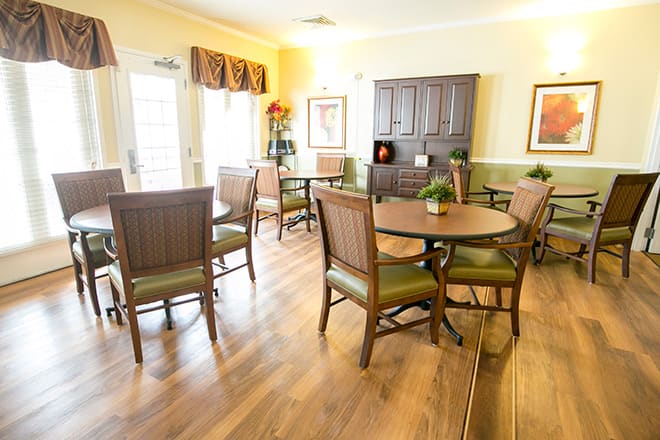 A bright dining room with several wooden tables and chairs, decorated with plants and artwork.