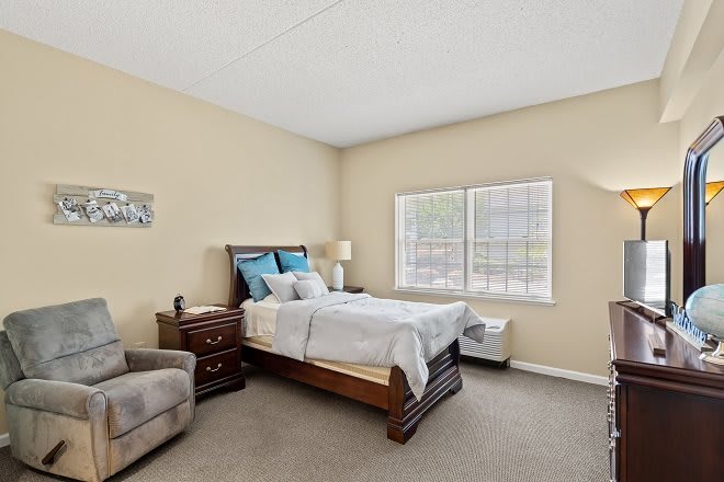 A cozy bedroom with a neatly made bed, a bedside table, and a comfortable chair by the window.