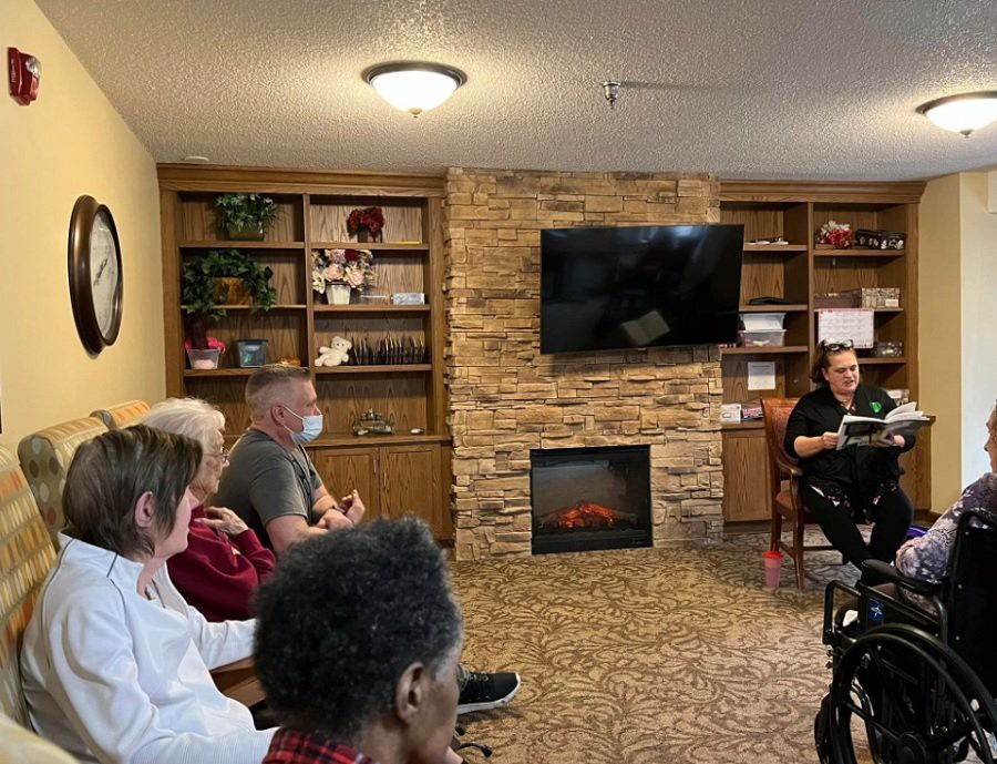 A group of people sitting in a cozy room with bookshelves and a fireplace, listening to a speaker.
