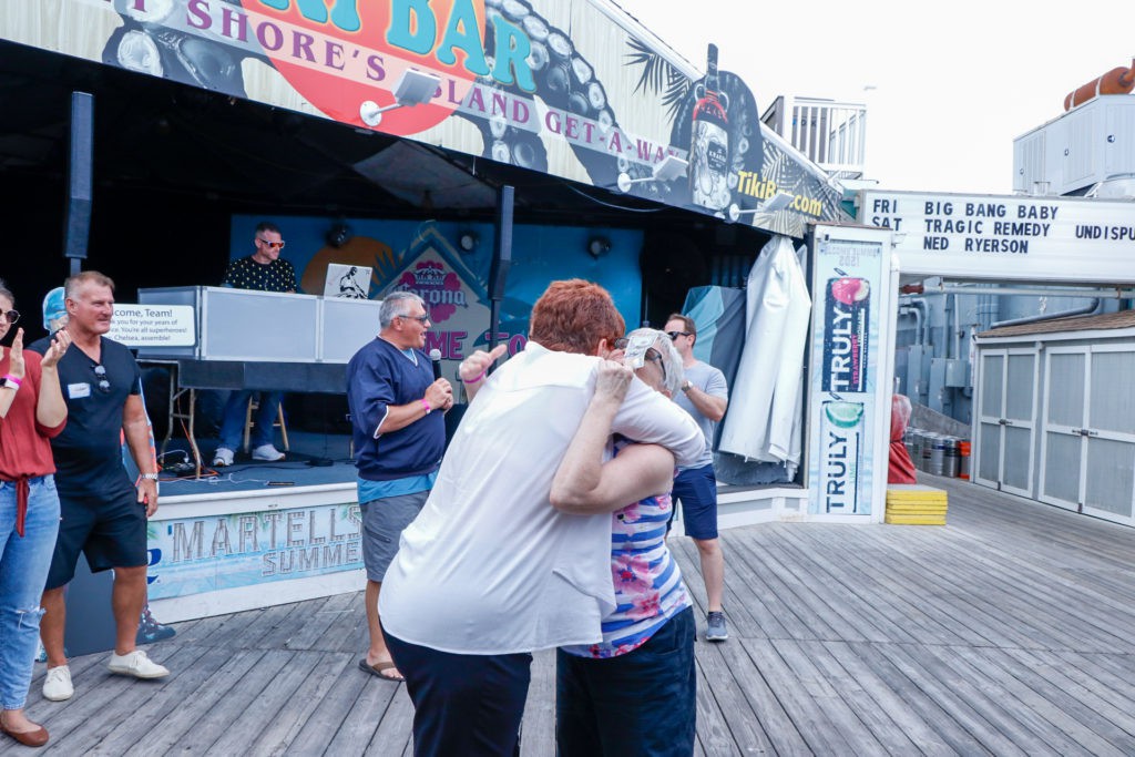 Two people share a hug on a boardwalk in front of a storefront.