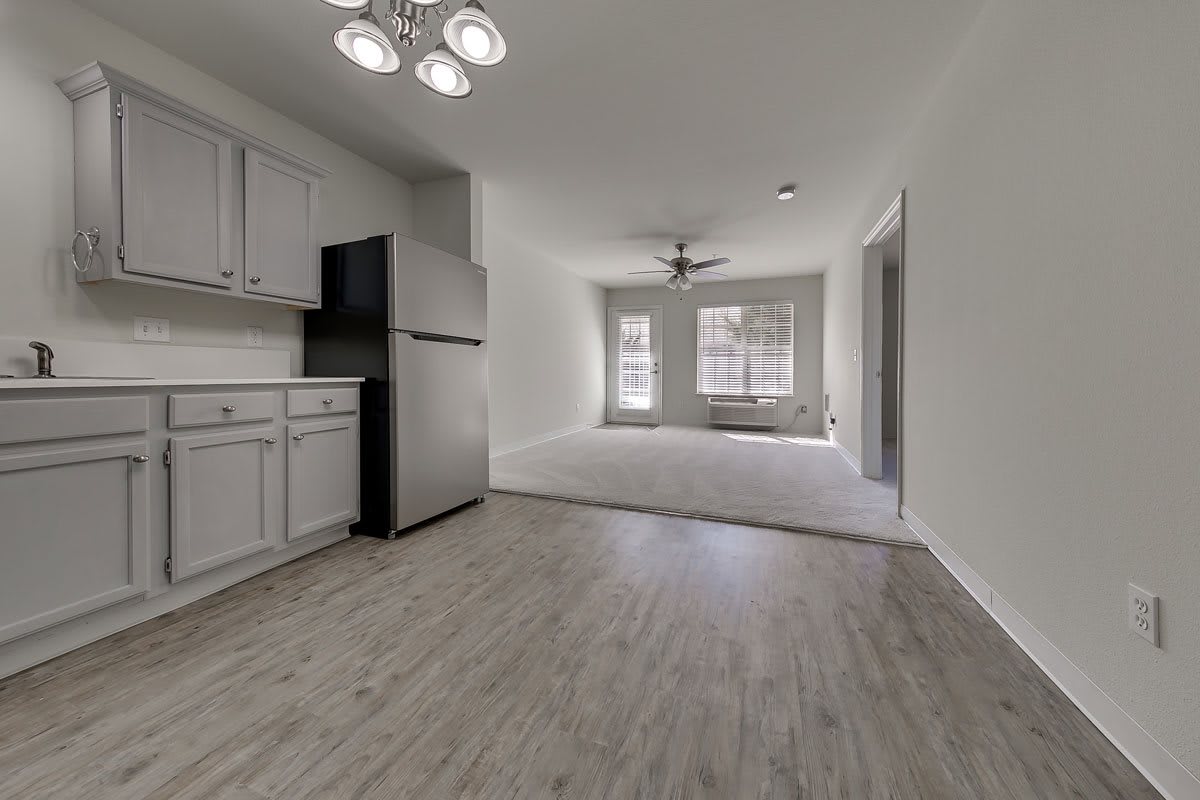 A modern apartment interior featuring a kitchen area with cabinets, fridge, and wood flooring adjacent to a living space with large windows and a ceiling fan.
