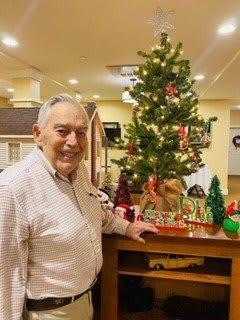 An elderly man standing beside a decorated Christmas tree in an indoor setting.