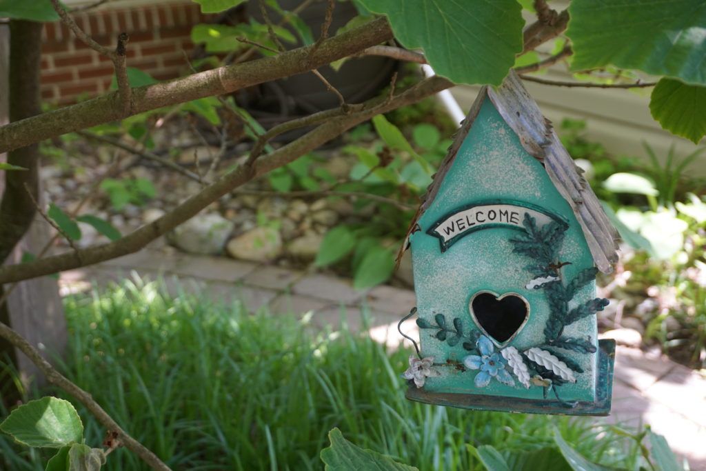 A decorative birdhouse with a welcome sign hanging in a lush garden.