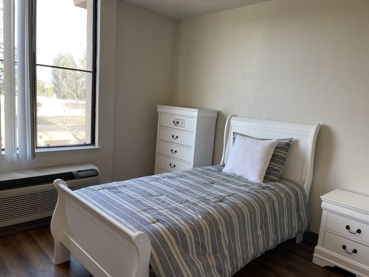 A minimalist bedroom with a single bed and dresser, featuring striped bedding and a large window.