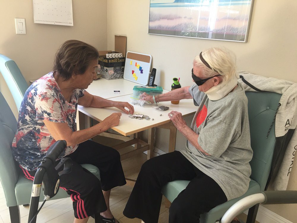 Two elderly women are sitting at a table working on a puzzle together indoors. One of them is wearing a mask.