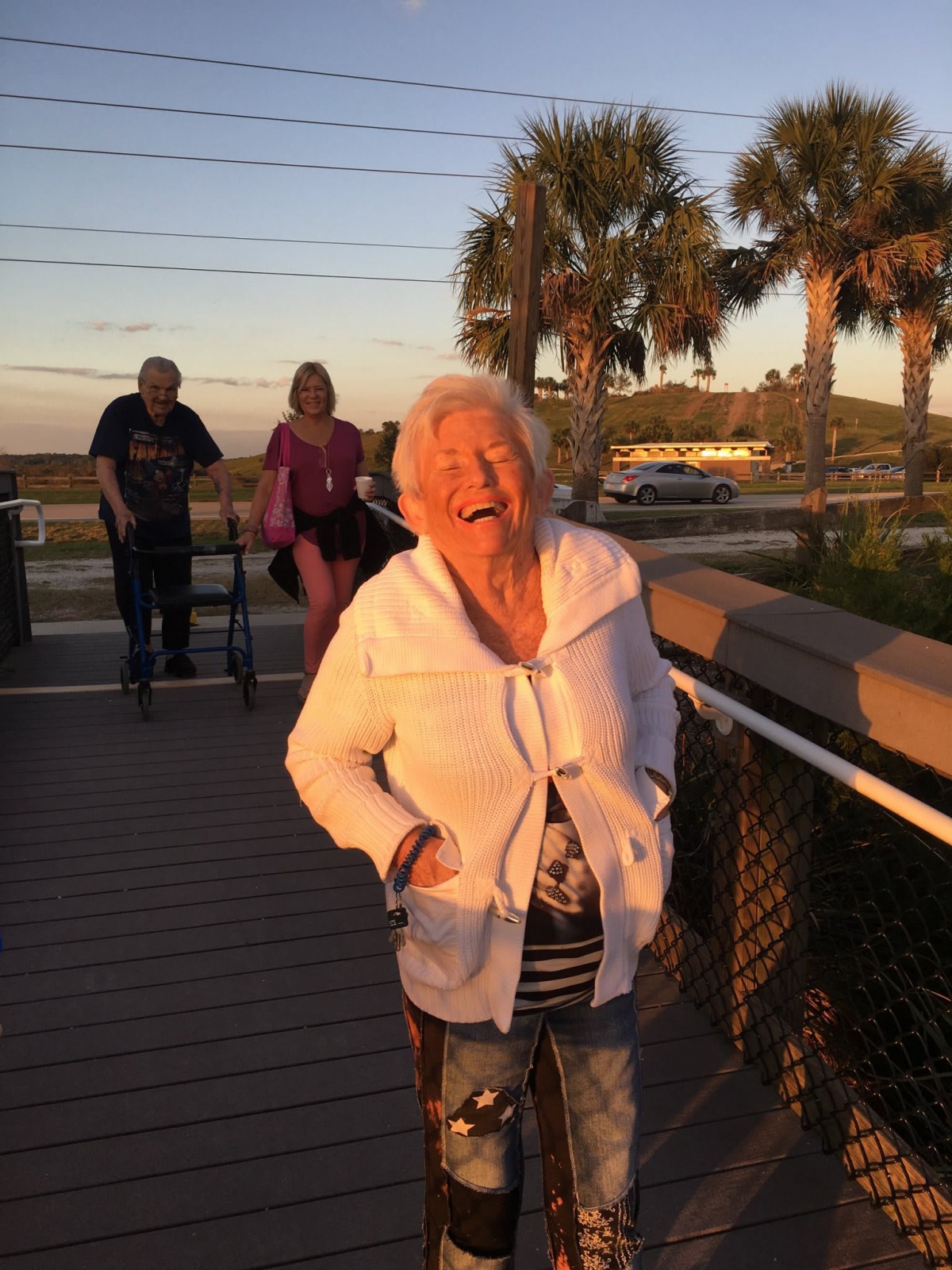 An elderly woman smiling joyfully while walking on a boardwalk during sunset.