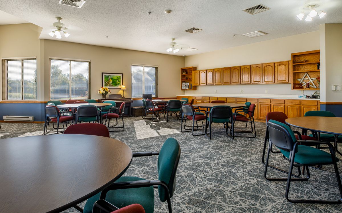 A spacious community room with tables and chairs, featuring a kitchenette and large windows.