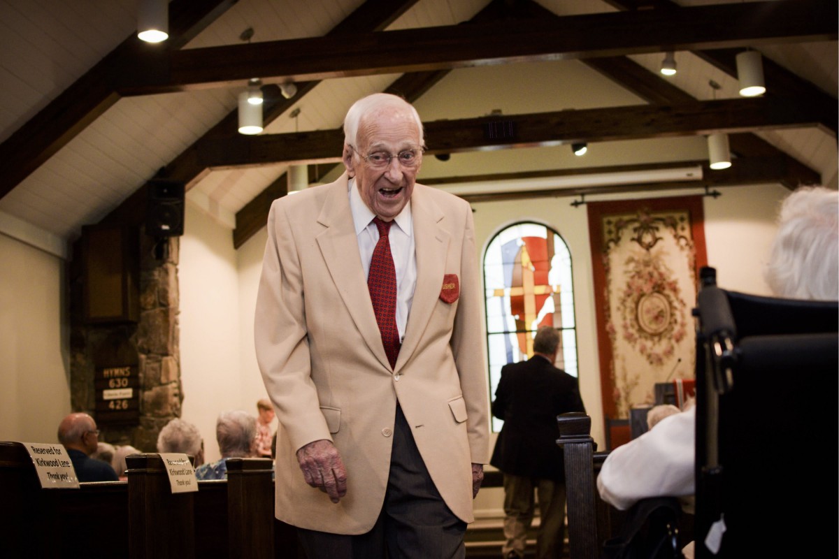 An elderly man in a beige suit and red tie walks inside a church during an event.