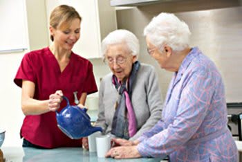 A nurse assisting two elderly women with pouring tea in a kitchen setting.
