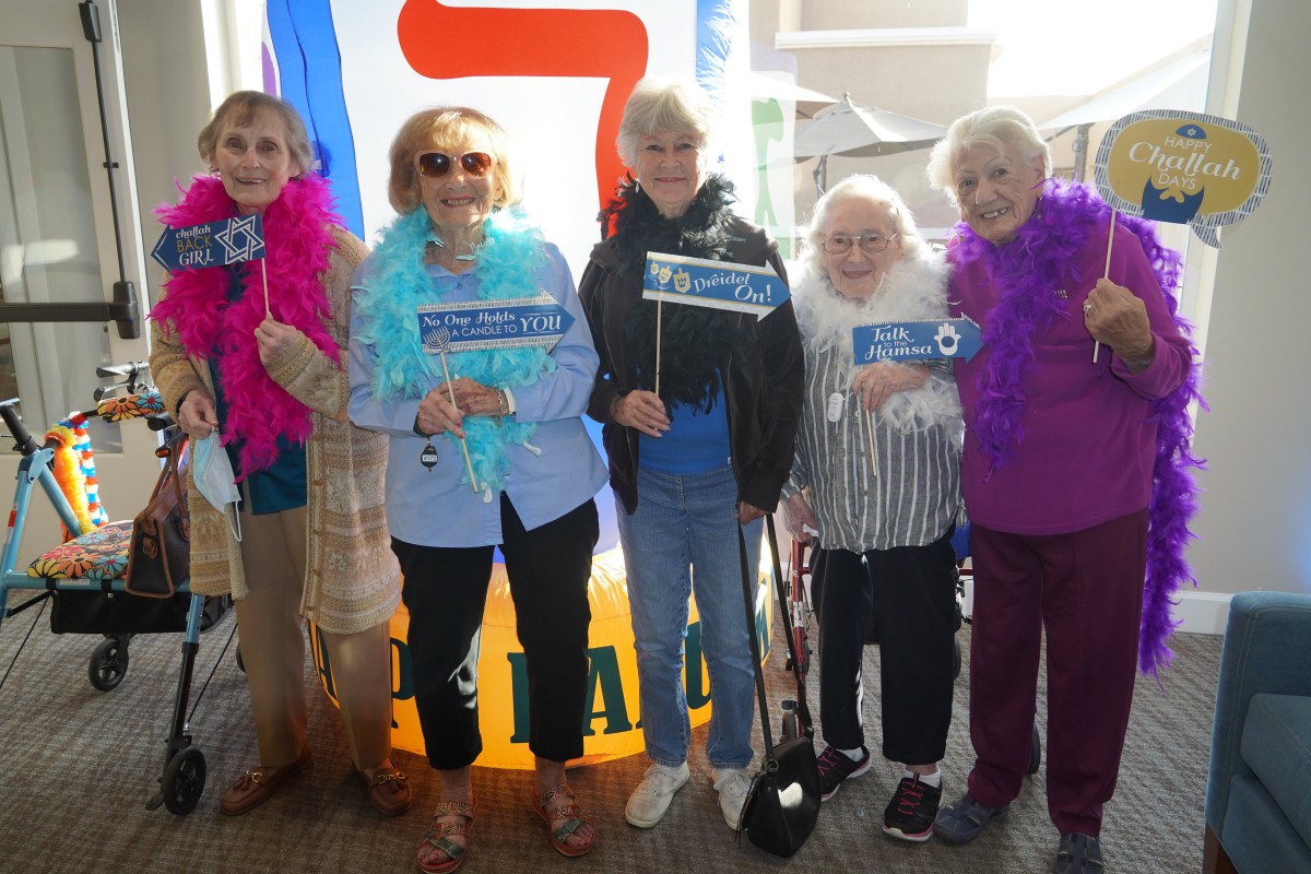 A group of older adults wearing feather boas, posing with signs and smiling indoors at a celebration event.