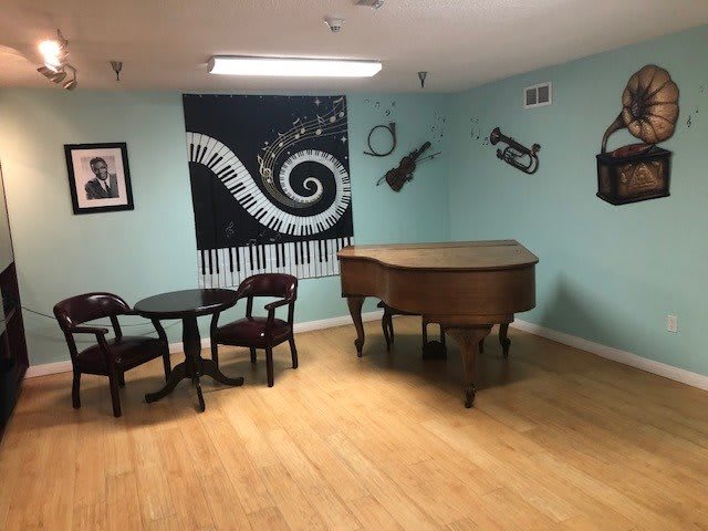 A cozy music-themed room featuring a grand piano, decorative wall art of musical instruments, and a small table with chairs on a hardwood floor.