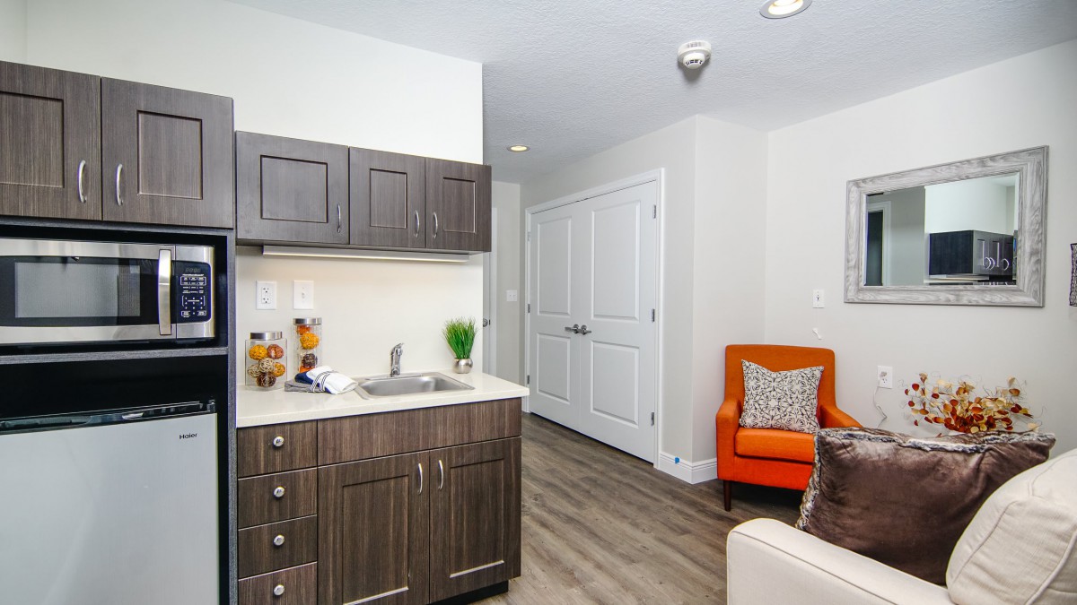 A modern kitchenette with dark cabinetry and an orange chair in the corner of a living space.