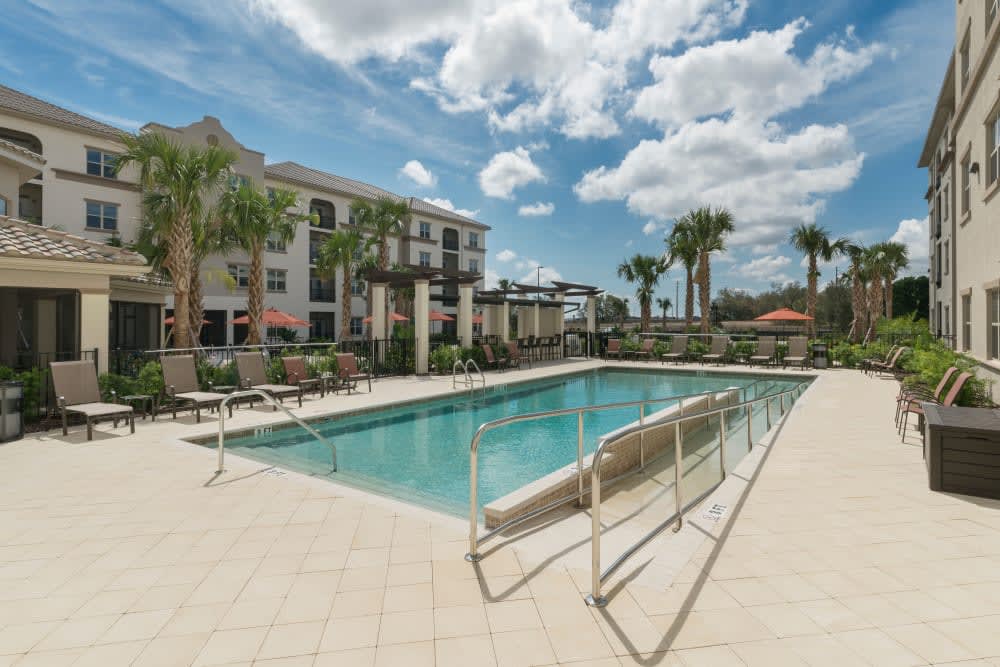 A swimming pool surrounded by a residential complex under a sunny sky with palm trees.