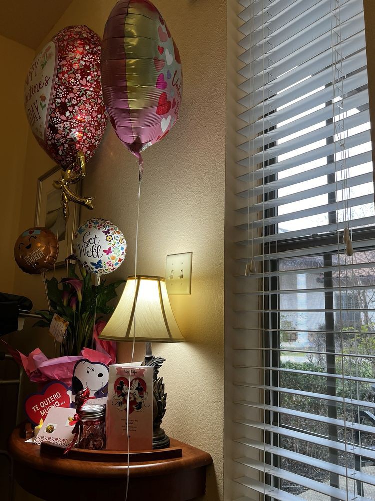 A cozy room corner with celebratory balloons, a bouquet of flowers, and a decorative lamp by the window.