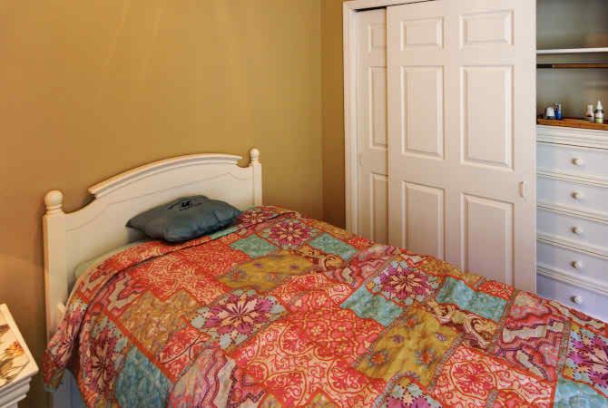 A cozy bedroom with a colorful quilt and a small pillow on the bed, set against a neutral wall with a white wardrobe.