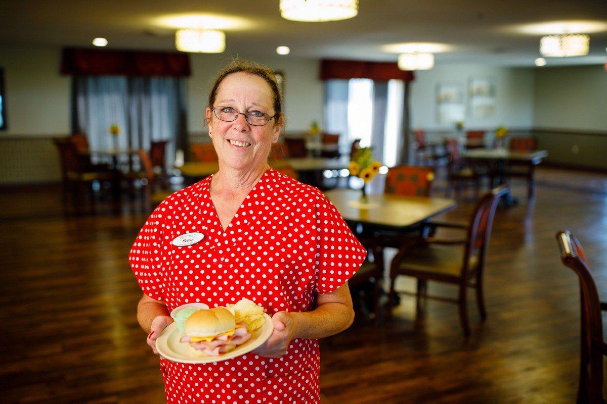 A smiling woman in a red polka dot shirt holds a plate inside a dining area.