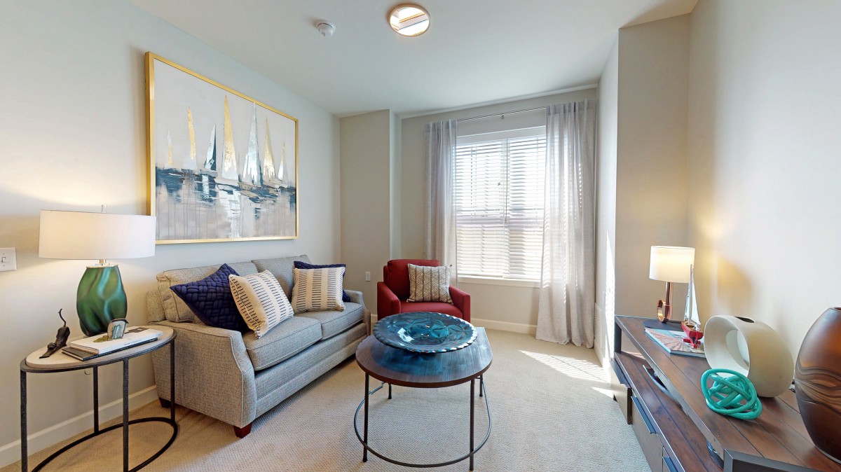 A cozy living room with a sofa and armchair, featuring a large window with curtains and a decorative painting on the wall.