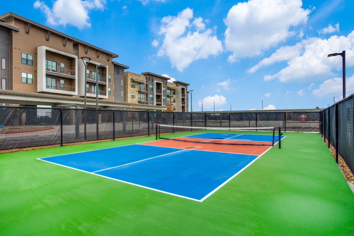 An outdoor tennis court with vibrant blue and green surfaces, surrounded by fencing and apartment buildings under a clear blue sky.
