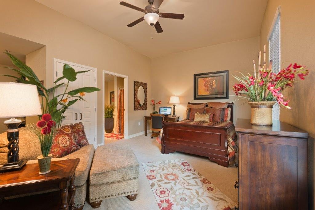 A cozy bedroom with traditional furnishings, featuring an armchair, a ceiling fan, and decorative plants.