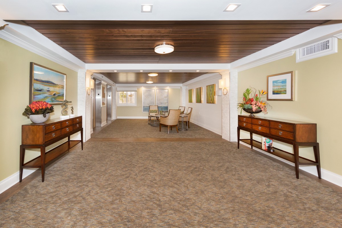 A spacious and elegantly decorated lobby area featuring a carpeted floor, wooden ceiling with lights, and furnishings including tables and chairs.