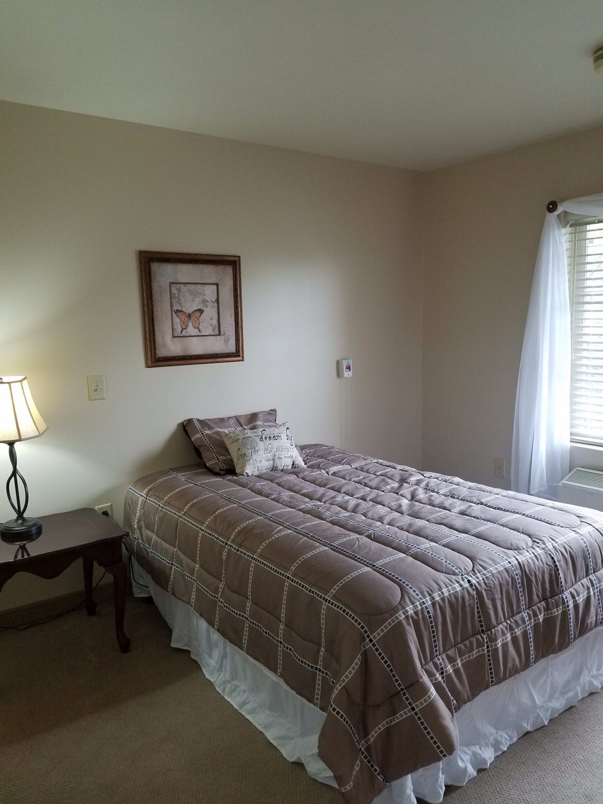 A cozy bedroom with a neatly made bed, a lamp on a side table, and a window letting in natural light.