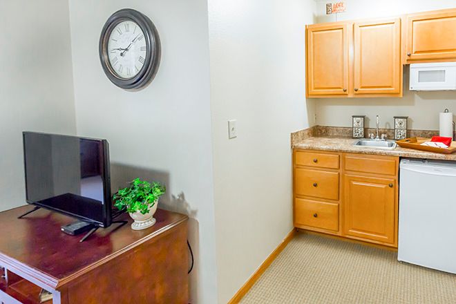 A small kitchenette area featuring wooden cabinets, a sink, a wall clock, and a nearby TV with a decorative plant.