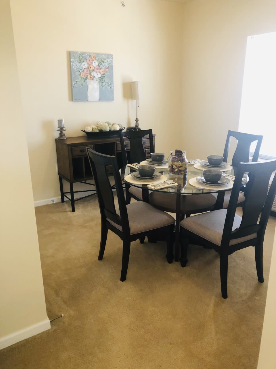 A cozy dining room with a round table set for four, featuring elegant decor and a sideboard with artwork above it.