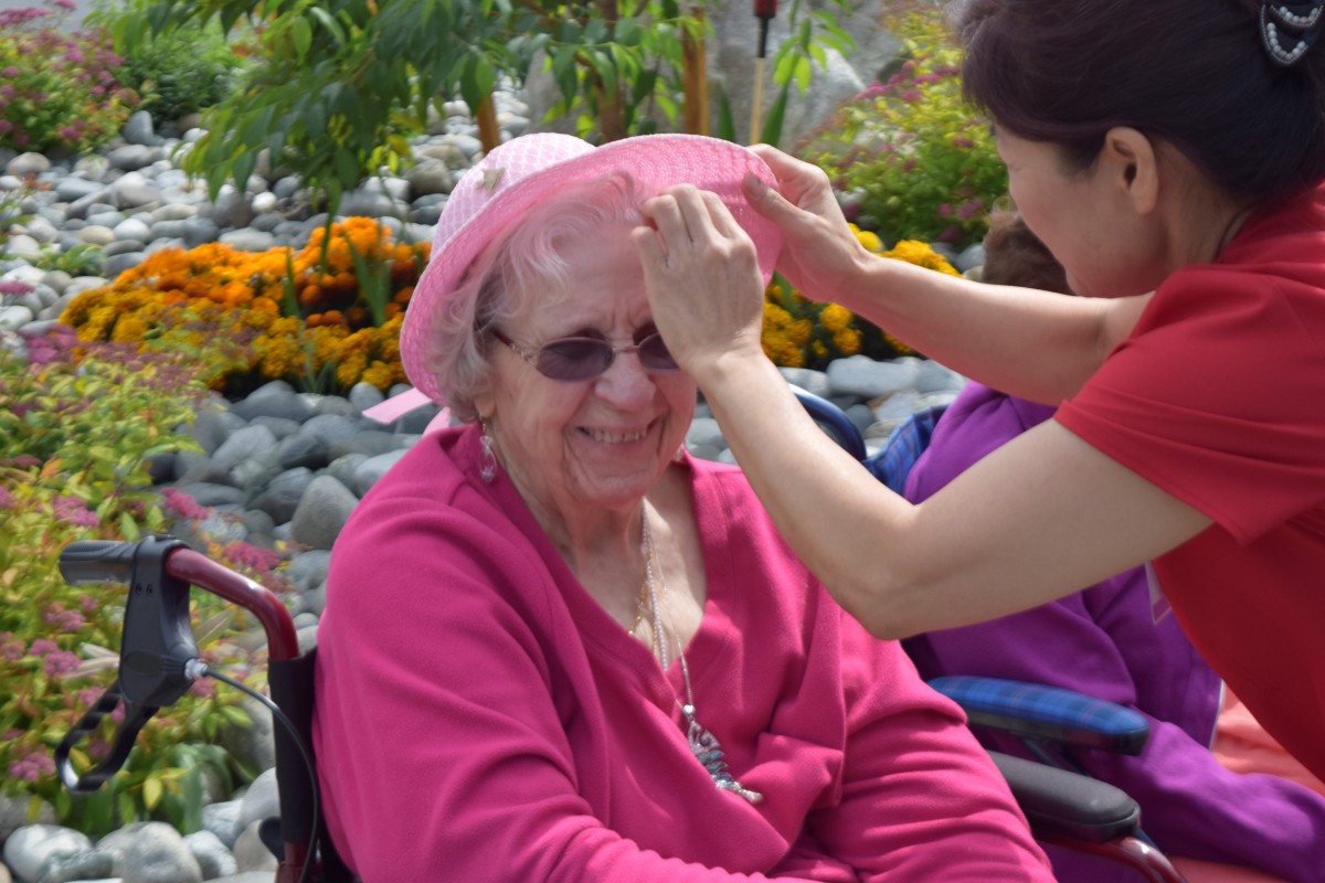 An elderly woman in a wheelchair enjoys a sunny day outdoors while someone adjusts her hat.