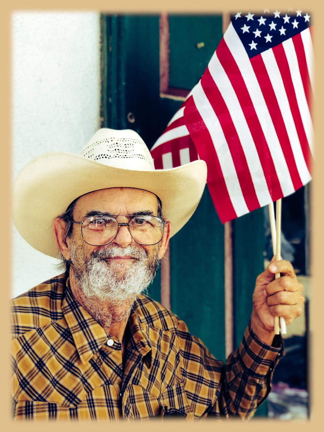 A smiling elderly man with a beard and glasses holds an American flag while wearing a cowboy hat and a plaid shirt.