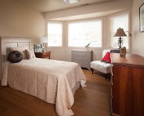 A cozy bedroom with a single bed, wooden furniture, and large windows letting in natural light.