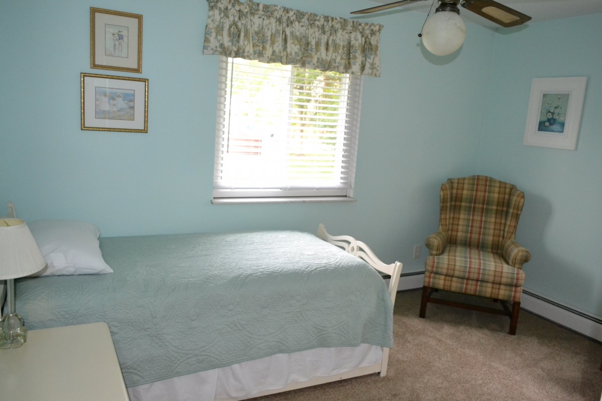 A cozy bedroom with a single bed and armchair, featuring light blue walls and a window bringing in natural light.