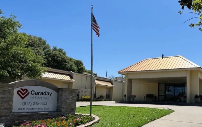 A building with an American flag in front, featuring a sign and surrounded by greenery under a clear blue sky.