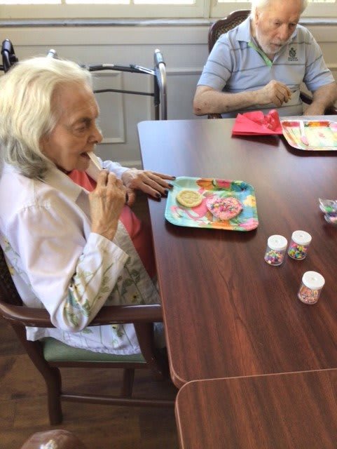 An elderly woman enjoys decorating cookies with sprinkles at a table.