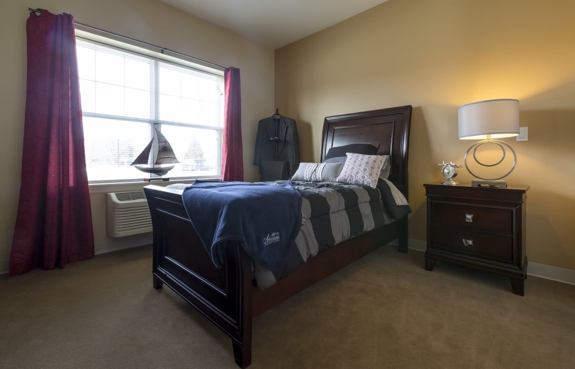 A cozy bedroom with a neatly made bed, lamp, and nightstand next to a large window.