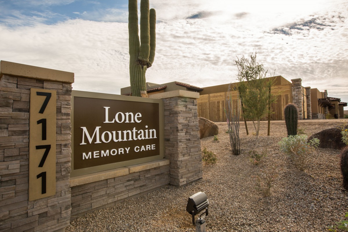 Entrance sign for Lone Mountain Memory Care set against a desert backdrop with a cactus and surrounding buildings.