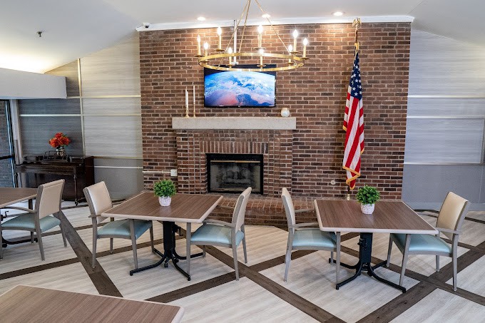 A cozy dining area with a brick fireplace, featuring modern tables and chairs, a TV mounted above the mantel, a chandelier overhead, and an American flag displayed in the corner.