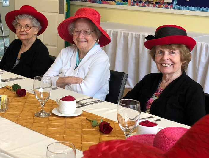 A group of elderly women wearing red hats enjoying a gathering at a table.