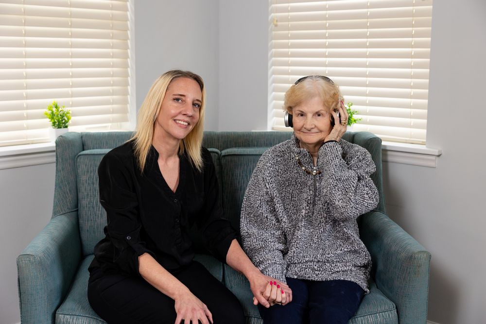 A younger woman and an elderly woman sit on a couch, smiling and holding hands. The elderly woman is wearing headphones.