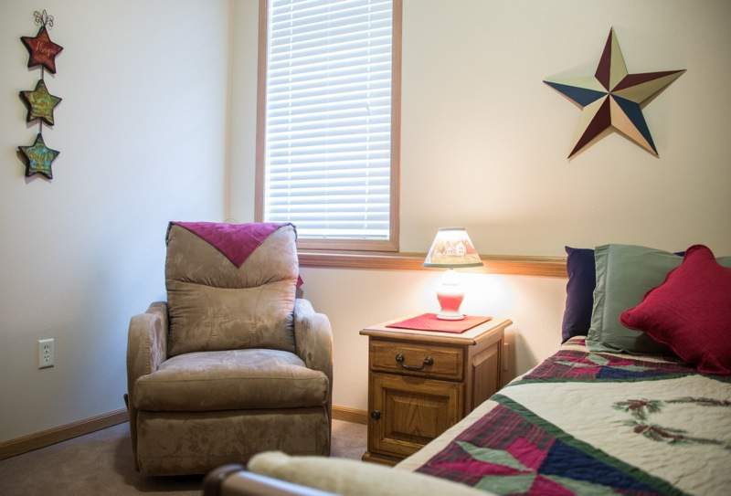 A cozy bedroom with an armchair, a bed adorned with a colorful quilt, and a wooden nightstand holding a lamp.