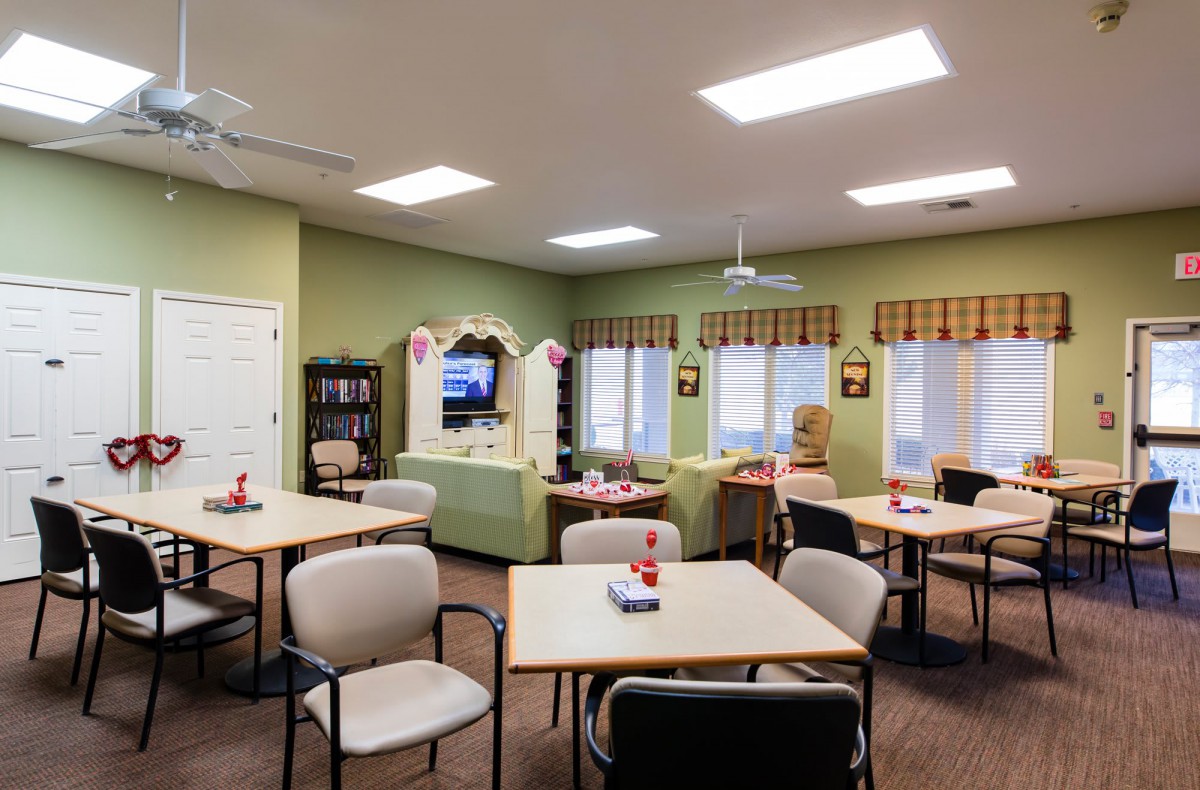 A cozy community room with tables, chairs, and a television, featuring green walls and natural light from multiple windows.