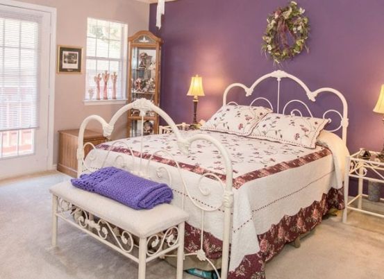 A cozy bedroom with a purple accent wall, featuring a white metal bed frame and floral bedding.