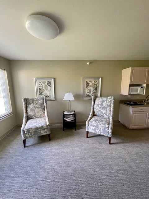 A minimalist living room featuring two patterned chairs, a small side table with a lamp, artwork on the walls, and a kitchenette in the corner.