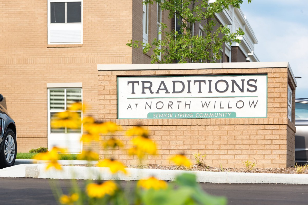 A sign for Traditions at North Willow in front of a building, with yellow flowers in the foreground.