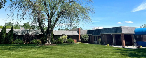 A brick house with a sprawling lawn and a large tree under a clear blue sky.